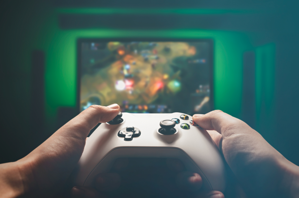 Intrepid Studios Class Action. View of gaming controller held by a gamer in foreground. In background, a computer game displayed on a big screen wall mounted TV.