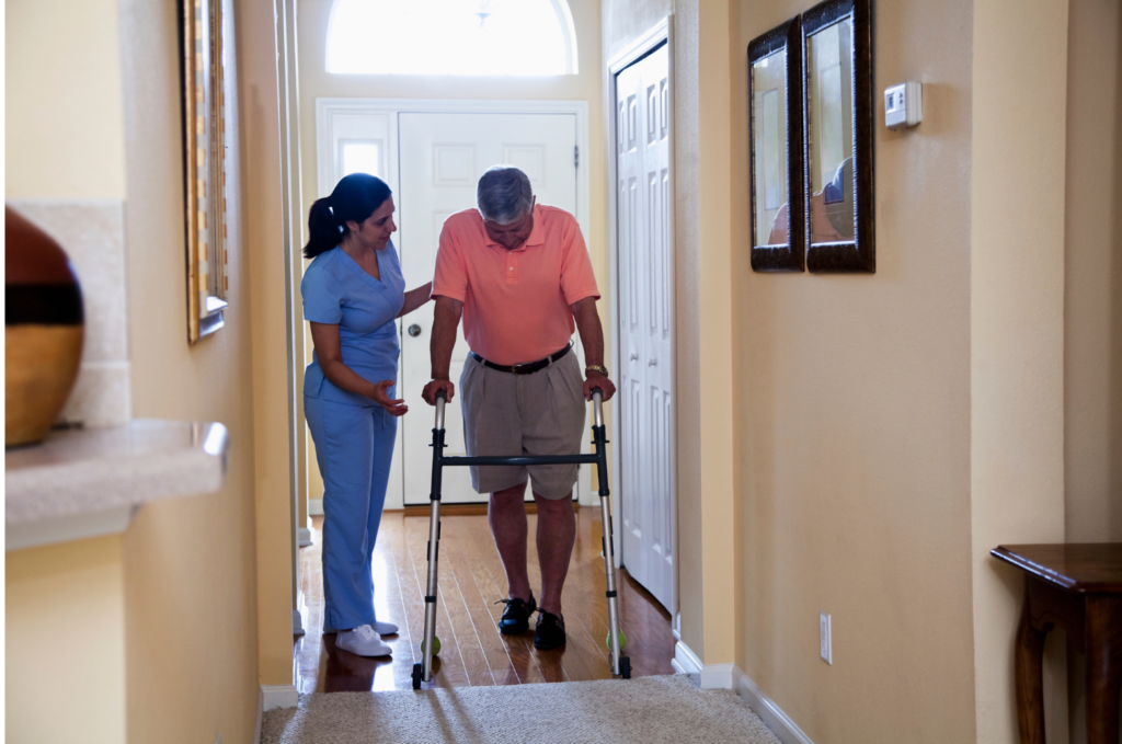 Anka WARN class action. Picture of a home health aide in blue scrubs helping a person in a pink polo shirt using a walker in a hallway.