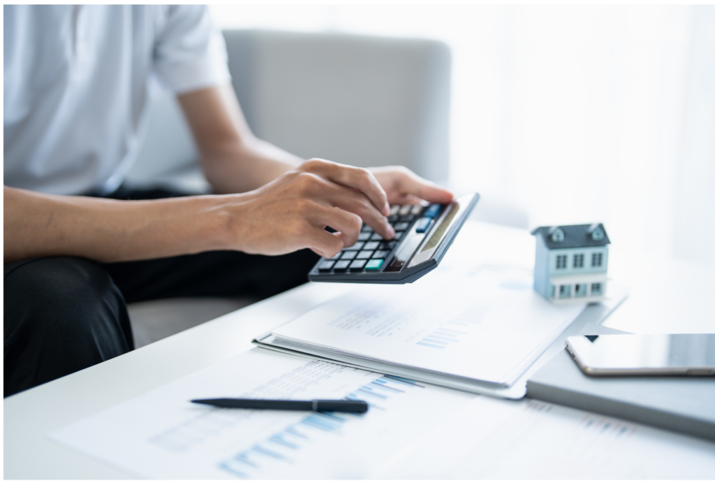 Live Well Financial WARN class action. View of person using a calculator to add up numbers for a potential mortgage. Financial forms and a small house shown slightly blurry in the background.