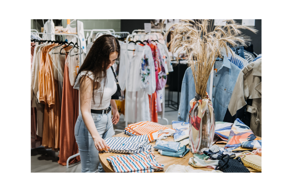 Avenue Stores WARN case. View of a woman shopping in a women's clothing store. She is looking at stacks of striped shirts on a table with racks of dresses behind her.