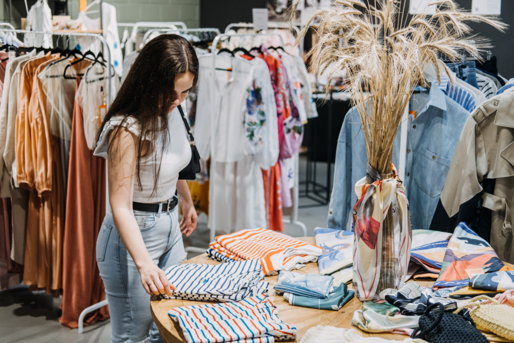 Avenue Stores WARN case. View of a woman shopping in a women's clothing store. She is looking at stacks of striped shirts on a table with racks of dresses behind her.