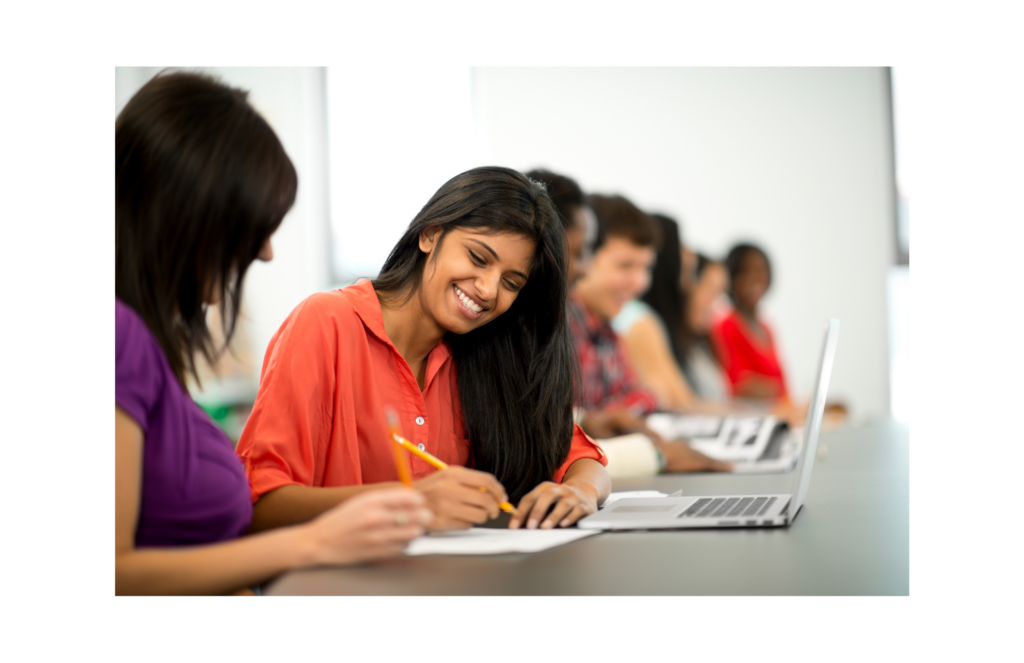CEHE WARN case. View of students at a table. In focus is a female student with a laptop, paper, and pencil who is discussing something with another female class mate.