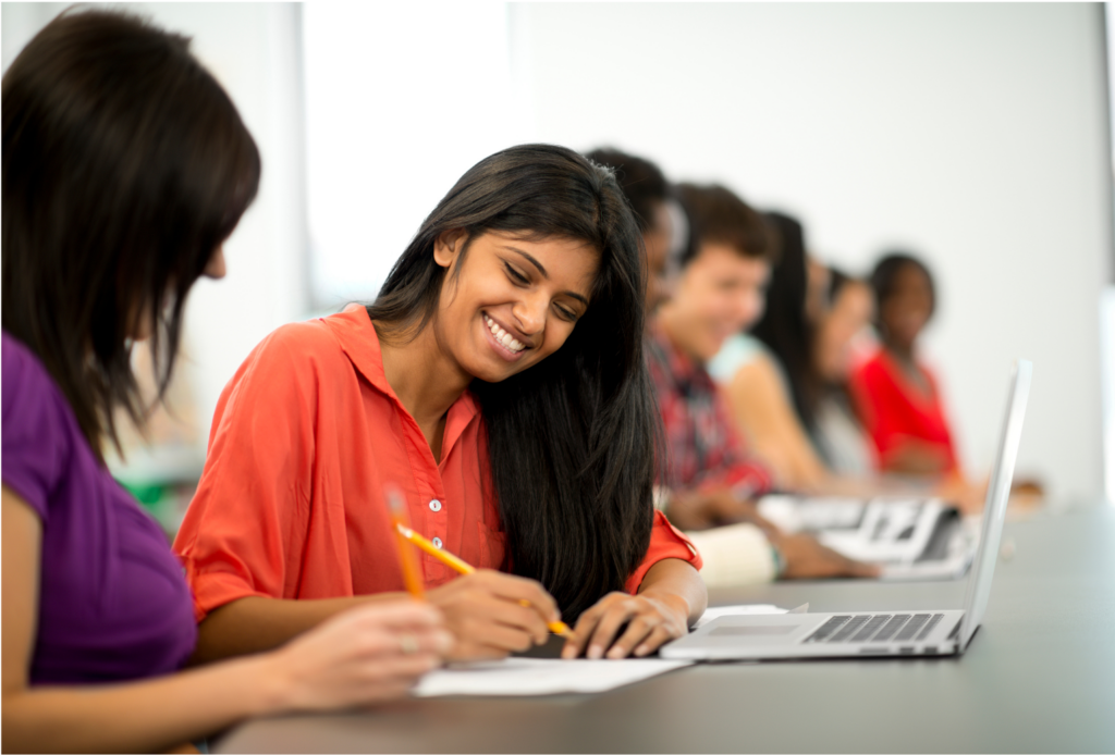CEHE WARN case. View of students at a table. In focus is a female student with a laptop, paper, and pencil who is discussing something with another female class mate.