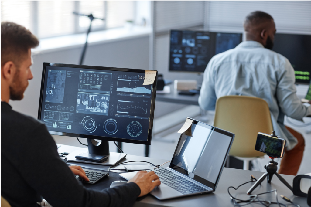GDIT WARN class action. View of two software developers, both using multiple computer screens, in a large modern office with bright sunlight and white walls.