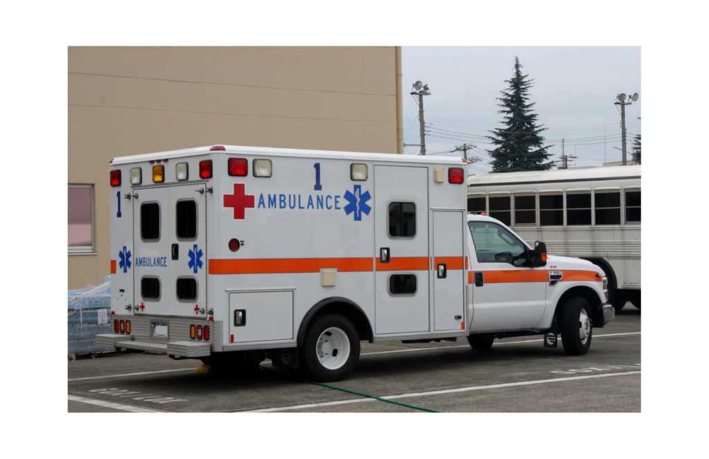Transcare WARN class action. View of an ambulance parked outside a municipal building with a bus painted in white in the background. The ambulance has a red cross and a blue rod of life--the symbol for ambulances in North America painted on its sides.
