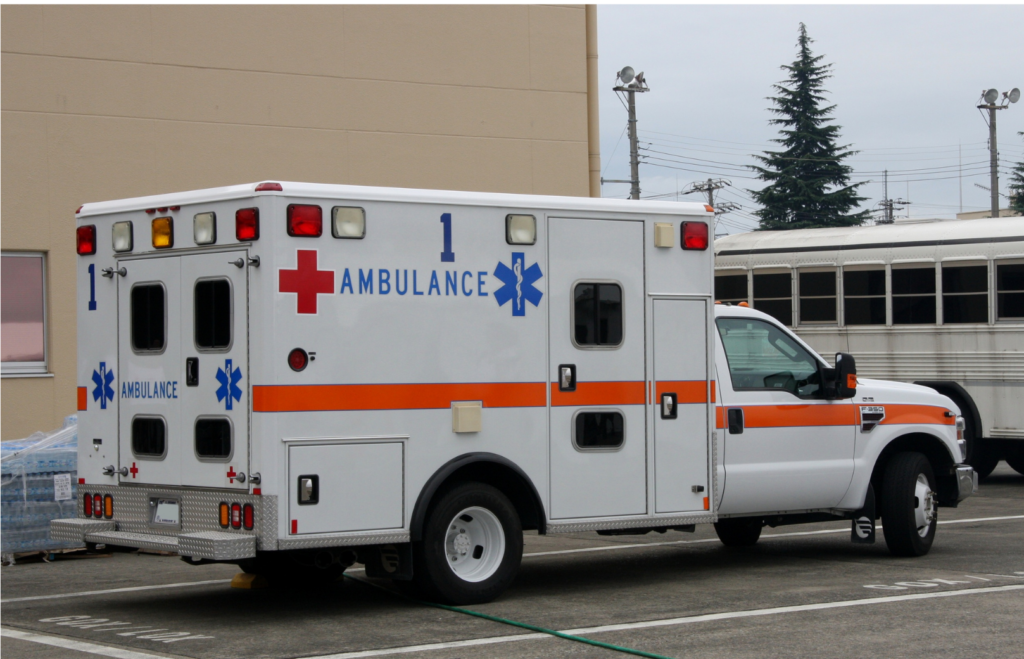 Transcare WARN class action. View of an ambulance parked outside a municipal building with a bus painted in white in the background. The ambulance has a red cross and a blue rod of life--the symbol for ambulances in North America painted on its sides.
