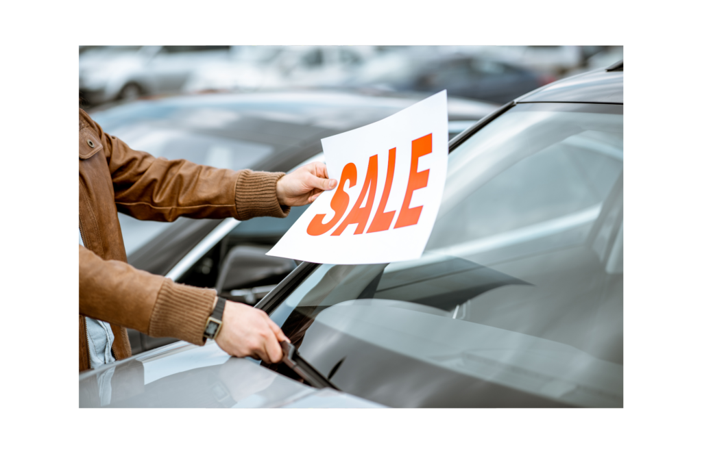 American Car Center WARN class action. View of a sales person putting a SALE sign on the windshield of a car in a small car sales lot. Blurred in the background are other cars for sale.