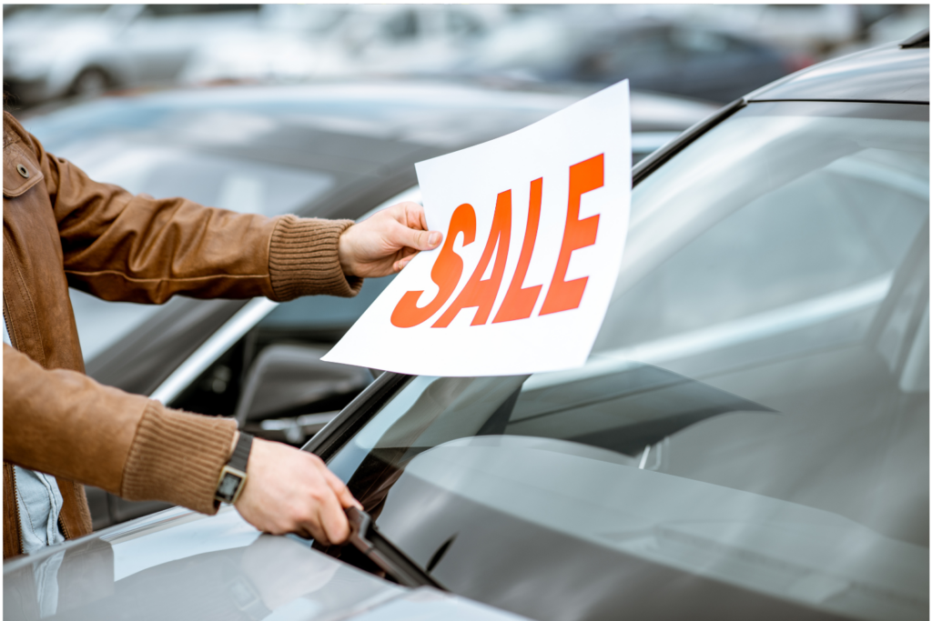 American Car Center WARN class action. View of a sales person putting a SALE sign on the windshield of a car in a small car sales lot. Blurred in the background are other cars for sale.