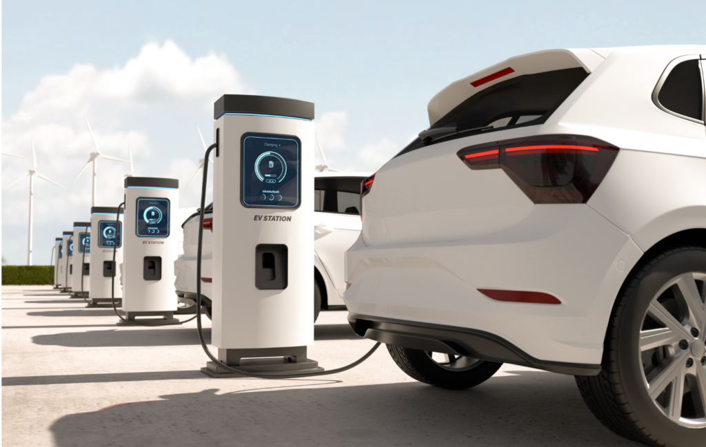 Canoo WARN class action. View of two new, white, electric vehicles at EV charging stations. In the background are numerous other charging stations and massive wind turbines in the distance.