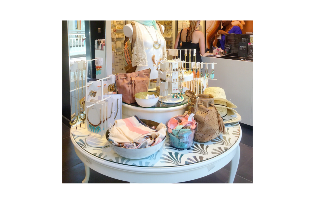 Charming Charlie WARN class action. View of a women's accessory store. A white table displaying hats, scarves, purses, and jewelry is shown in the foreground. Behind that, a woman is paying for her purchase at a sales counter. Along the wall of the store are numerous earrings and necklaces for sale.