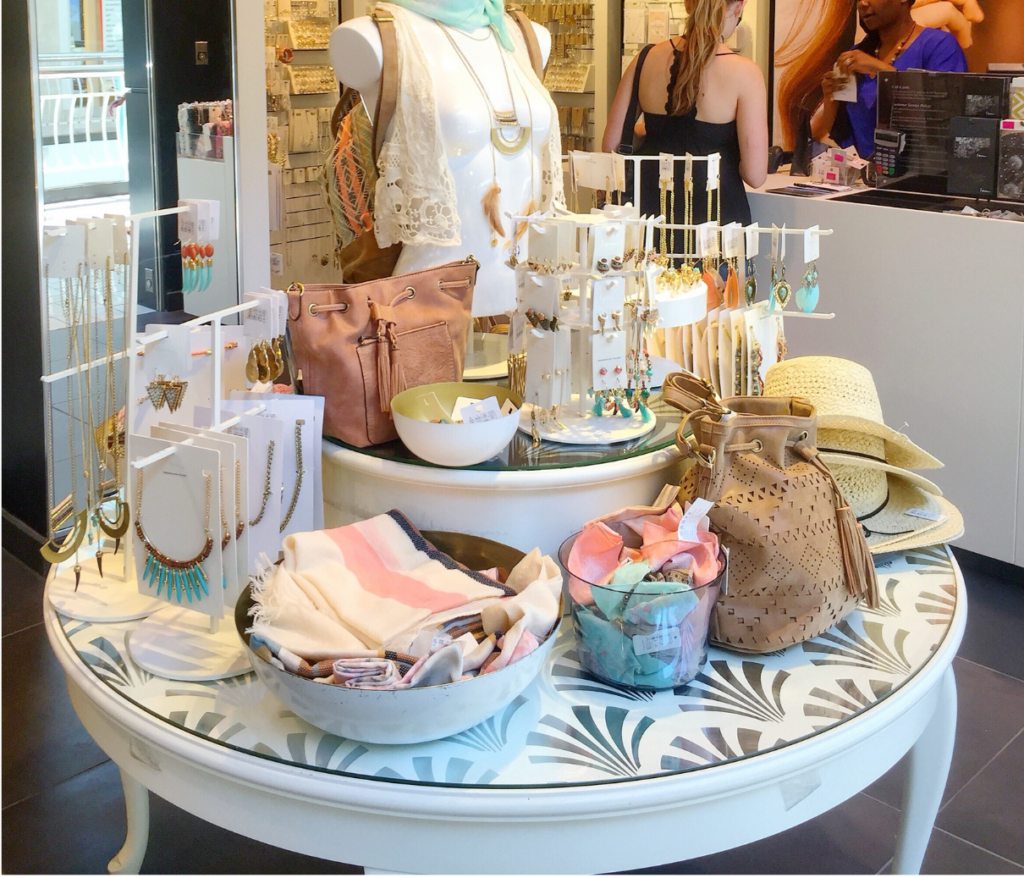 Charming Charlie WARN class action. View of a women's accessory store. A white table displaying hats, scarves, purses, and jewelry is shown in the foreground. Behind that, a woman is paying for her purchase at a sales counter. Along the wall of the store are numerous earrings and necklaces for sale.
