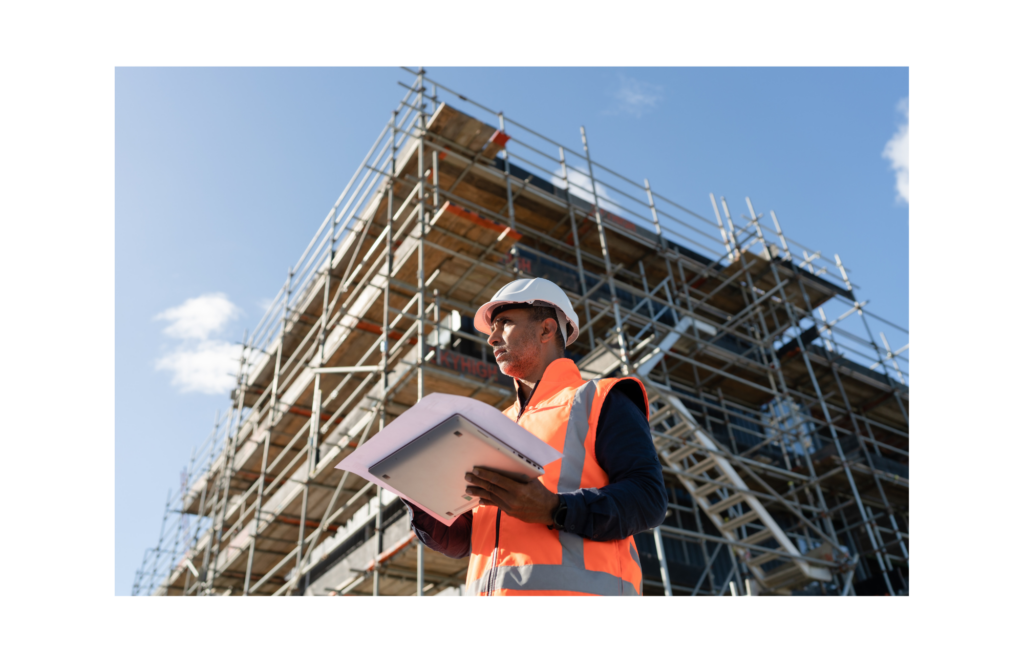 Commodore WARN class action. View of a construction manager wearing an orange vest and a white hard hat and holding a laptop and blue prints in front of a multi-story building being built.