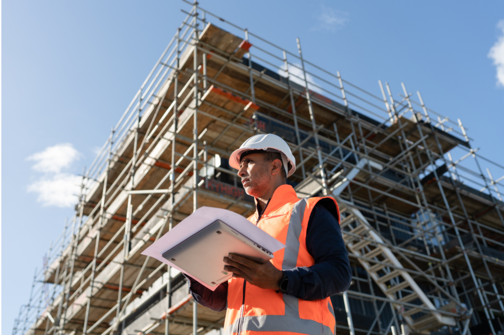 Commodore WARN class action. View of a construction manager wearing an orange vest and a white hard hat and holding a laptop and blue prints in front of a multi-story building being built.