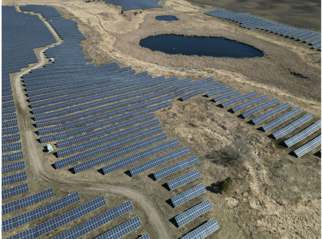 DC Solar WARN class action. View of a large solar field with the solar panels looking somewhat like a jigsaw puzzle. The panels are built in a dry field with scrubby vegetation. There are a couple of small ponds amidst the solar field of panels.