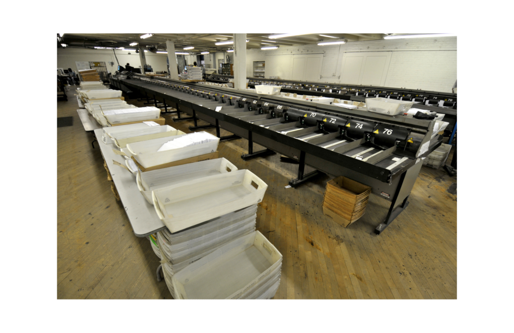 DGI Services WARN class action. View of a large empty mail sorting room with numerous bins filled or containing rows of envelopes. The room is white with a wooden floor and hanging fluorescent lights on the ceiling.