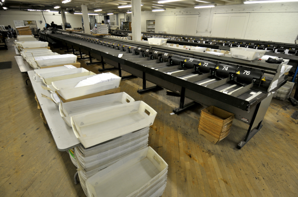 DGI Services WARN class action. View of a large empty mail sorting room with numerous bins filled or containing rows of envelopes. The room is white with a wooden floor and hanging fluorescent lights on the ceiling.
