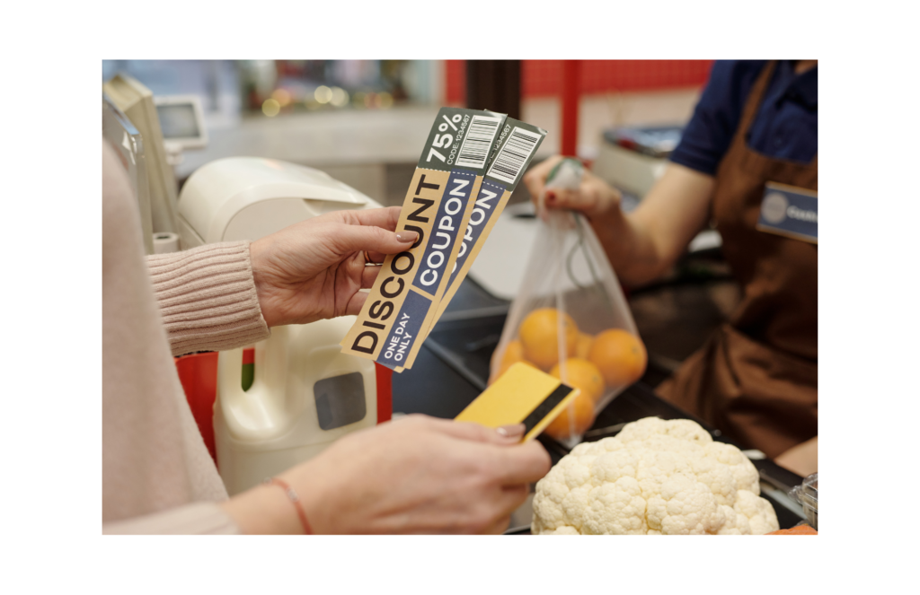 Entertainment Publications WARN class action. View of a woman in a pale peach colored sweater using 75% off large coupons to pay for cauliflower, oranges and milk in a market. The person working the register is shown blurred out in the background.