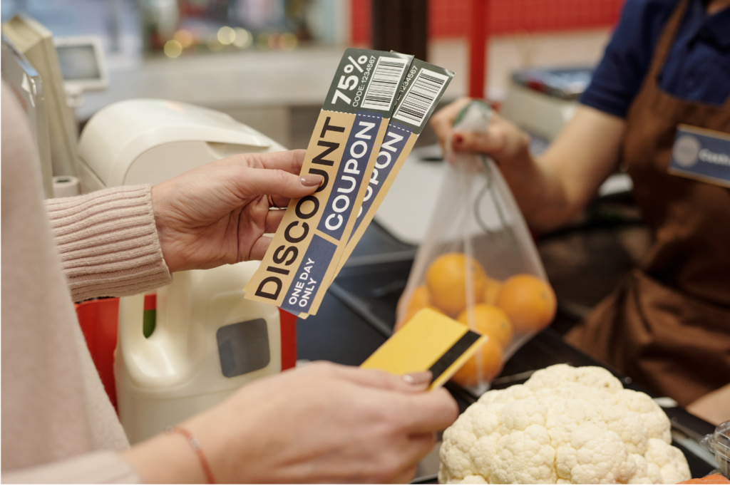 Entertainment Publications WARN class action. View of a woman in a pale peach colored sweater using 75% off large coupons to pay for cauliflower, oranges and milk in a market. The person working the register is shown blurred out in the background.