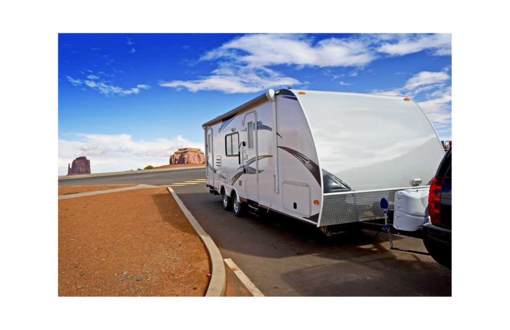Evergreen Recreational Vehicles WARN class action. View of a white, towed, sleeper trailer hitched behind an SUV. The SUV and trailer are parked beside a curb but the background is very austere brown dirt with high rocky towers in the background.