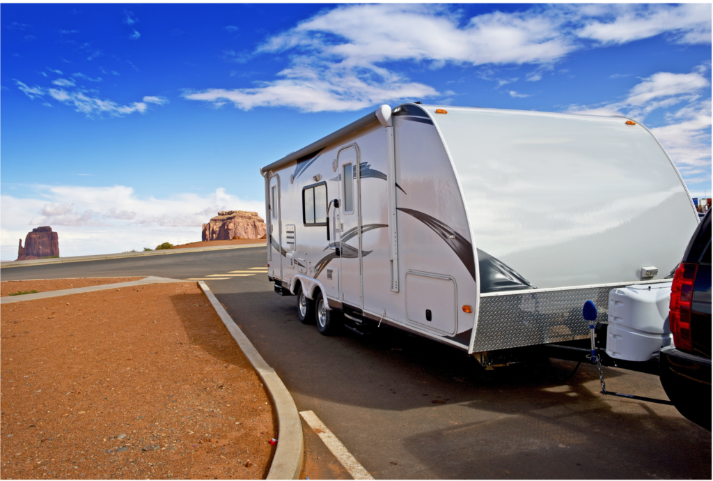 Evergreen Recreational Vehicles WARN class action. View of a white, towed, sleeper trailer hitched behind an SUV. The SUV and trailer are parked beside a curb but the background is very austere brown dirt with high rocky towers in the background.