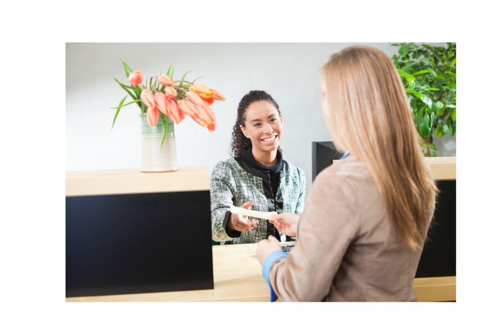 First Guaranty Mortgage Corporation WARN class action. View of a bank teller accepting a deposit from a woman customer. There are flowers on the bank counter and a tree in against the white wall in the background.