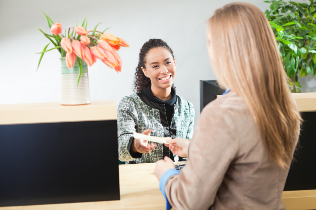 First Guaranty Mortgage Corporation WARN class action. View of a bank teller accepting a deposit from a woman customer. There are flowers on the bank counter and a tree in against the white wall in the background.