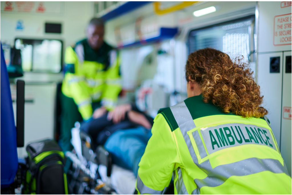 FirstMed EMS WARN class action. View of two ambulance workers wearing neon green/yellow jackets with forest green and gray reflective markings putting a patient into an ambulance on a gurney. The first responder closest to the camera is in focus. Otherwise everything else is blurry.