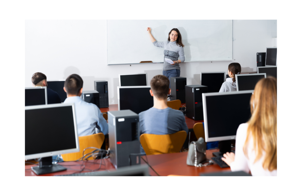 ITT Educational Services WARN class action. View of a cramped smaller classroom with several students in rows sitting in front of desktop computers. The teacher is a casually dressed woman who is standing in front of a wide whiteboard with no writing.
