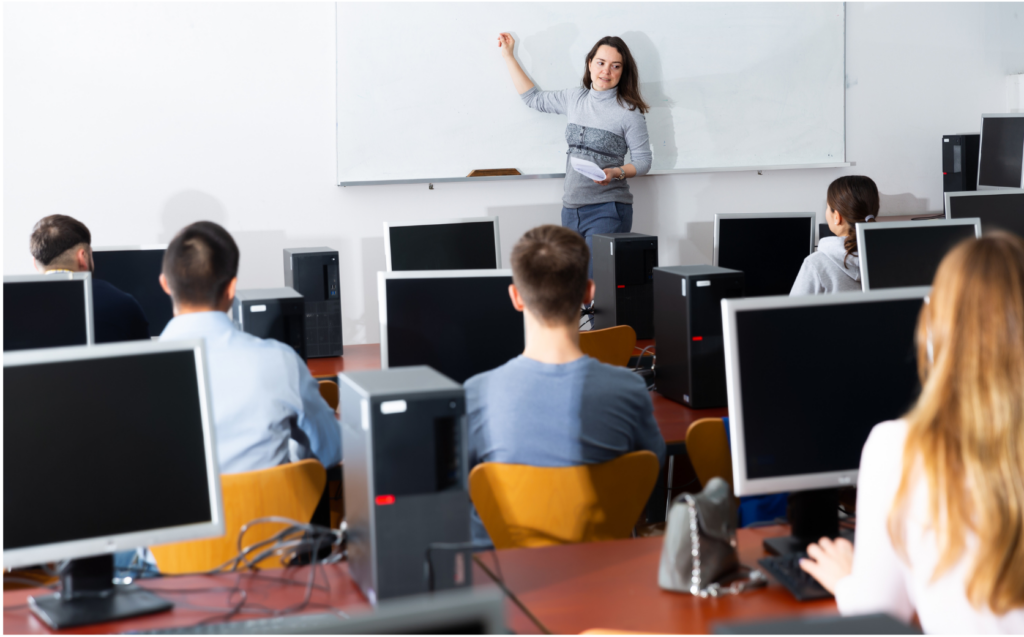ITT Educational Services WARN class action. View of a cramped smaller classroom with several students in rows sitting in front of desktop computers. The teacher is a casually dressed woman who is standing in front of a wide whiteboard with no writing.