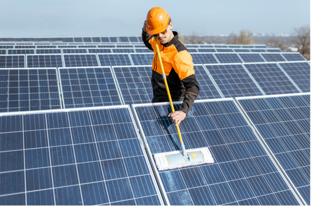Meyer Burger WARN class action. View of a worker in orange and black and with a black hard hat using a swiffer type mop to clean solar panels. He is standing amidst an entire field of solar panels.