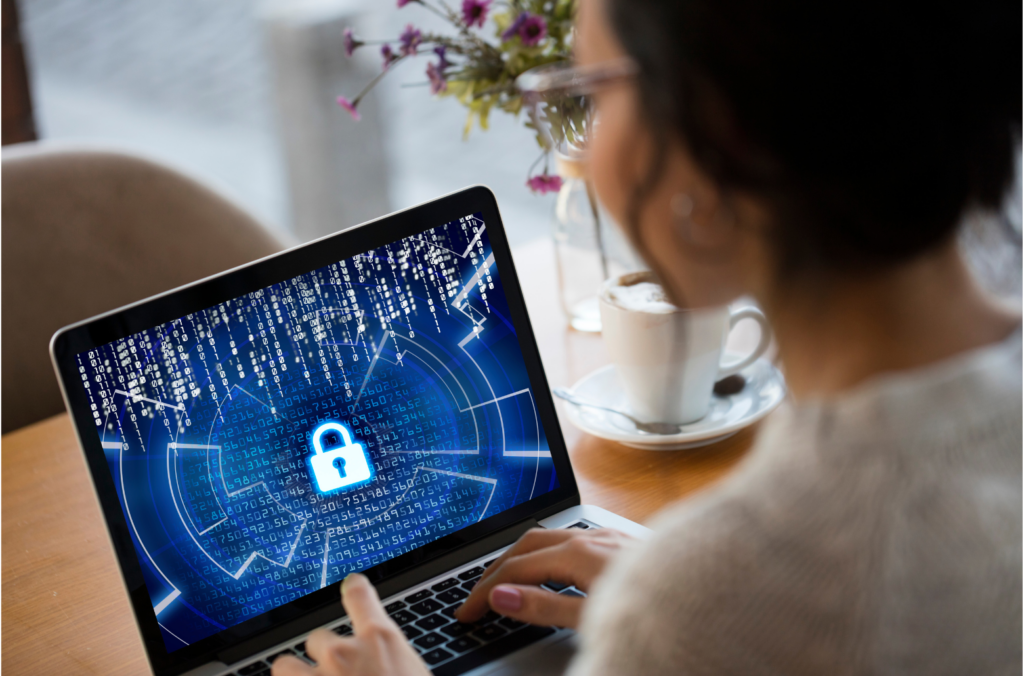 NS8 WARN class action. A woman with glasses using a laptop at her desk. Flowers and a cup and saucer are on the desk. The laptop displays a centered padlock icon with a grid of random numerals displayed in white against a blue background.