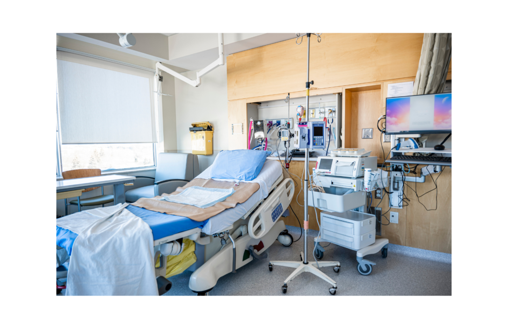 Santa Cruz Valley Regional Hospital WARN class action. View of a modern hospital room with a hospital bed, iv stand, ekg machine on a cart, numerous tubes and outlets on the wall to attach addition equipment. There is a desk and chair in the room in front of windows and a computer screen and keyboard on a stand extended from the wall behind the head of the bed.
