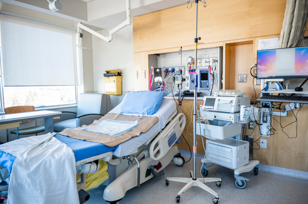 Santa Cruz Valley Regional Hospital WARN class action. View of a modern hospital room with a hospital bed, iv stand, ekg machine on a cart, numerous tubes and outlets on the wall to attach addition equipment. There is a desk and chair in the room in front of windows and a computer screen and keyboard on a stand extended from the wall behind the head of the bed.