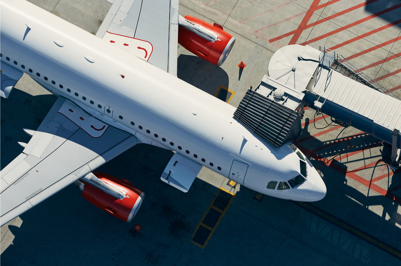 Overhead view of a large multi-engine commercial airliner at the gate of a modern airport. The walkway for passengers is extended to meet the aircraft.