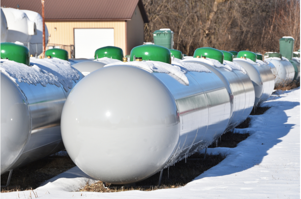Worley & Obetz | Amerigreen Propane WARN class action. View of rows of gray large propane tanks with green caps. Each tank appears to be 8-10 feet wide and four feet high. There is snow on the ground and the tops of the propane tanks. Buildings and trees are shows in the background.